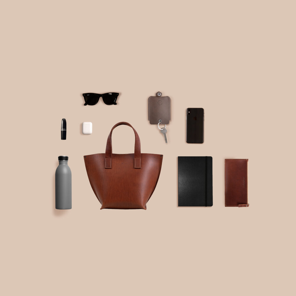 A Model Bucket - Cognac, sunglasses, and other accessories laid out on a beige surface.