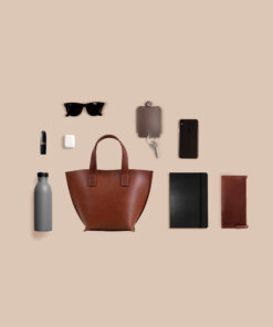 Un bolso de cuero marrón, gafas de sol y otros accesorios dispuestos sobre una superficie beige.